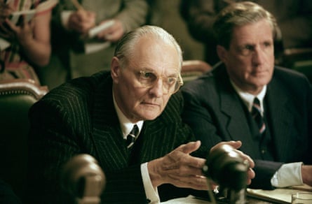 Alan Alda balding in black suit and tie leaning forward to a microphone - with another suited man to his left