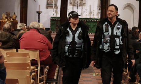 Two police officers walk in the aisle of a church with people sitting in chairs on either side