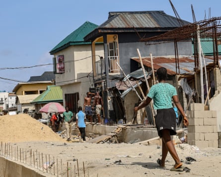 People walk past a half-built wall and a pile of sand next to houses in a street