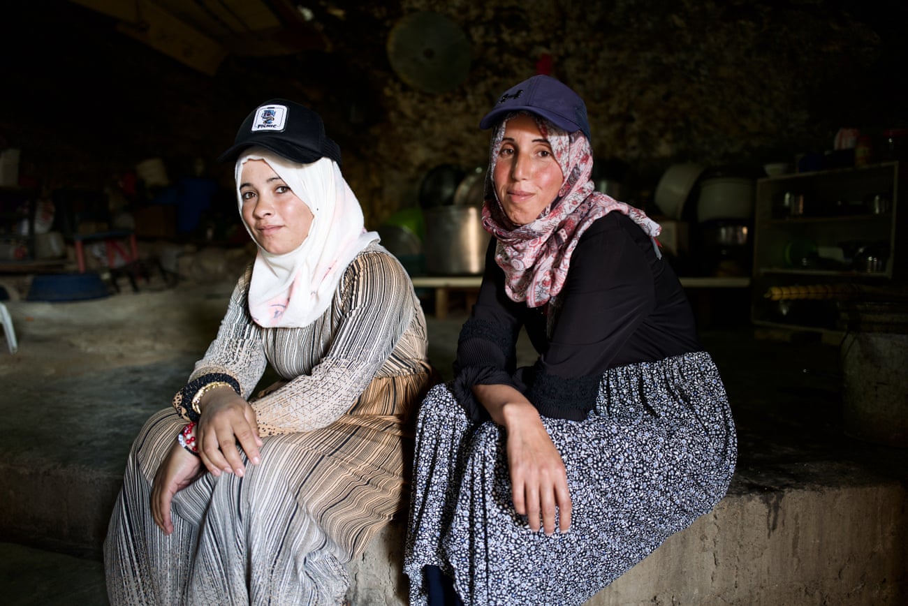 Two younger women sit and give the camera wry smiles. Both wear baseball caps over their headscarves