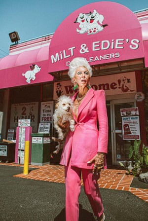 A slim, tanned, bouffant elderly lady in a bright fuchsia suit (low-cut jacket, no blouse) emerges from a fuchsia shop with bouffant lapdog under her arm