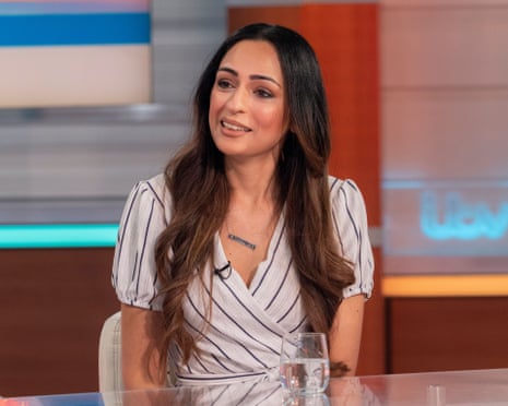 Bushra Shaikh sits at a desk wearing a striped wrap dress on a television set
