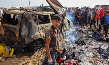 A boy with cuts on his face and a distressed look on his face stands in front of a burned car and debris from the explosion at the camp