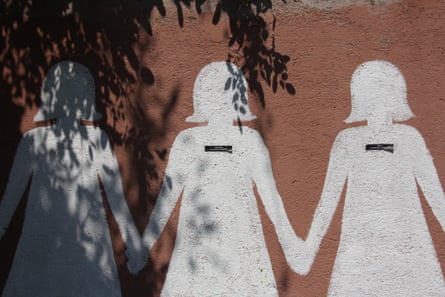 A mural on a red wall of faceless women painted in the style of a chain of paper dolls with black rectangular stickers on their chests giving a woman’s name and a date.