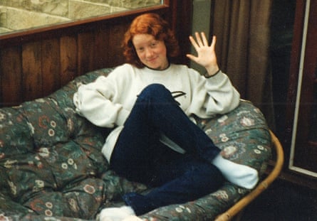 A red-haired teenage girl smiling shyly and waving at the camera reclined on a patterned sofa – wearing white sweatshirt and black leggings and white socks