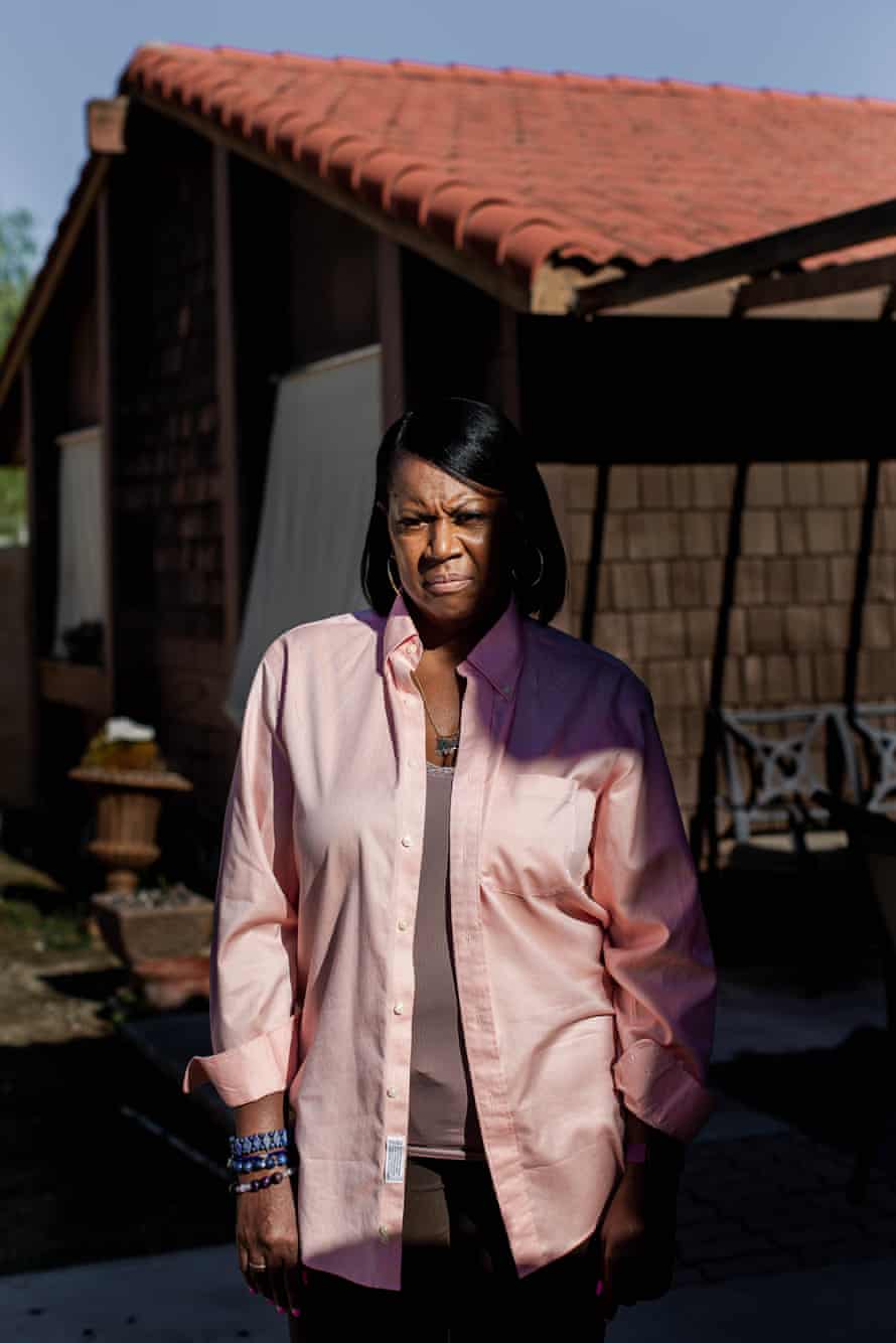 A black woman wearing an open salmon button down poses for a portrait in front of her home