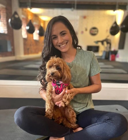 Leanne Lucas in a yoga or exercise studio; she is sitting cross-legged on a mat holding a small brown dog with curly fur and a bandana tied around its neck; she has long dark hair and wears a loose light green T-shirt and dark leggings, and is smiling in this photo.