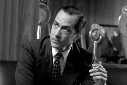 A black-and-white still of a man in a suit holding an old-timey microphone.