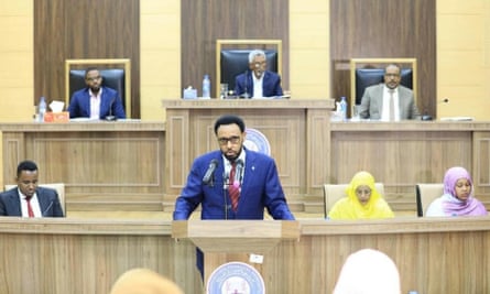 A man wearing a blue suit stands at a lectern with government officials seated behind him.