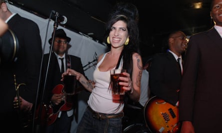 Amy Winehouse smiles as she stands on stage holding a drink in each hand, with musicians behind her