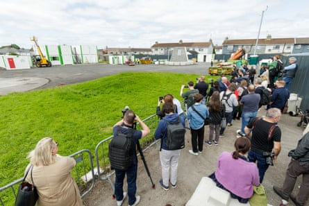 a group of people stand behind temporary railings by a grassy rectangle with houses to the side.