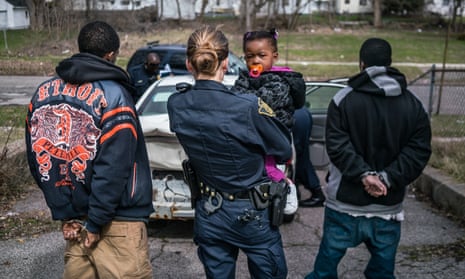 An officer holds a small child while her father is being searched and questioned.