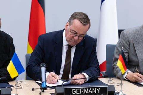 Germany’s defence minister, Boris Pistorius, signs a document at a meeting with ‘Germany’ sign in front of him and flags