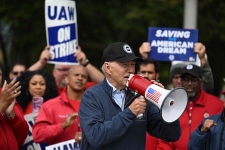 Joe Biden, wearing a navy blue zip-up and a black United Auto Workers baseball cap, speaks into a megaphone at a union strike.