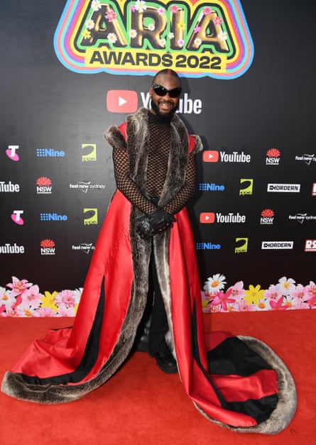 Genesis Owusu on the red carpet wearing a red fur lined cape and black mesh top and black sunglasses