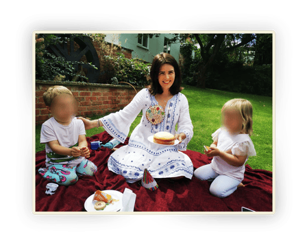 Matilda Davis holds a cake while she sits next to two children, their faces blurred out, on a picnic rug in the garden of a house