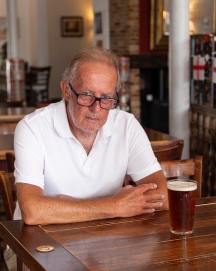 A man wearing glasses and a white polo shirt sits at a polished wooden table in a pub, with a pint of bitter