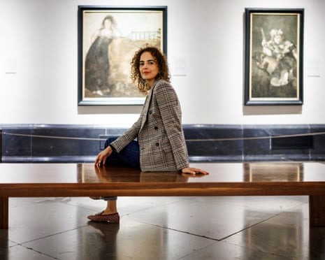 Writer Leila Slimani sits on a wooden bench in a gallery, wearing a checked blazer