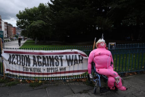 A pink blown up mannequin depicting Trump on a wheelchair next to a banner reading 'Action against war'