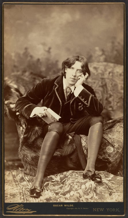 Oscar Wilde, 1882, is photographed in a plush jacket sitting on a large upholstered chair and with knee-high socks and fancy shoes