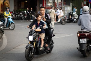 A child sitting up front while a man drives a motorbike and checks his phone