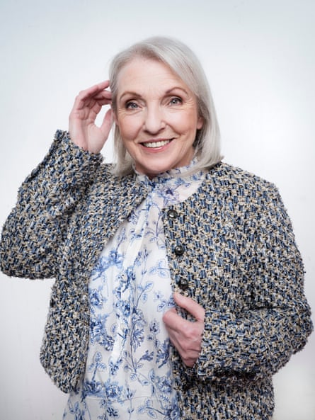 A woman with short grey blond hair, wearing a black speckled jacket and a blue printed shirt.
