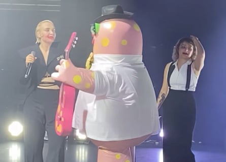 An inflatable pink creature with yellow spots on stage with two musicians