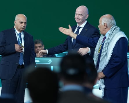 Head of Israel’s football federation, Basim Sheikh Suliman, Fifa boss, Gianni Infantino and Jibril Rajoub in suits stand on a stage at a football congress. Jibril Rajoub, wearing a black-and-white keffiyeh scarf, gestures across the stage.