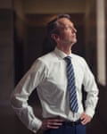 Andy Haldane stands in a dark corridor with the light focused on him; he has his hands on his hips and his face is turned almost in profile. He wears a bright white shirt and dark striped tie.