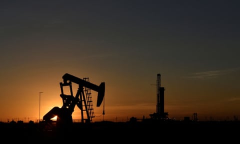 A pump jack operates in front of a drilling rig at sunset in an oil field in Texas, US.