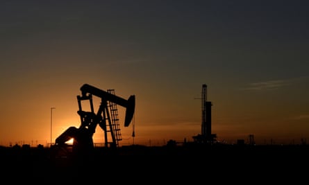 A pump jack operates in front of a drilling rig at sunset in an oilfield in Midland, Texas.