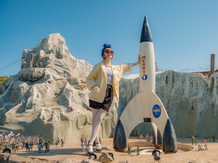 A woman with bright orange hair, leaning against a model rocket, with rocks behind her and tiny people below, while wearing sunglasses, a big blue bow in her hair, a yellow striped jacket, a blue striped top and red lipstick
