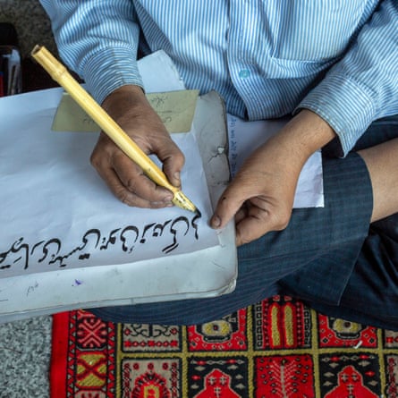 A man sits cross-legged writing with a large pen made of bamboo.