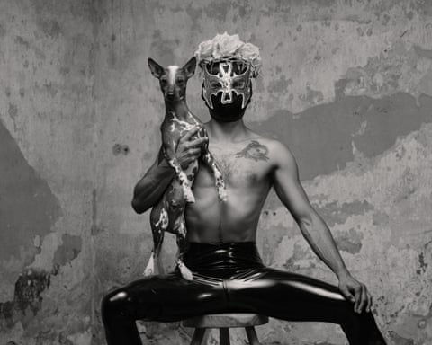 A shirtless person in a wrestling mask and leather pants sits holding a spotted dog