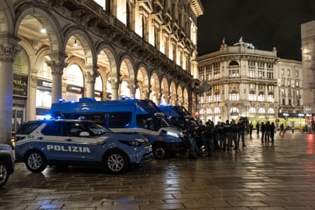 Blue and white police vehicles are lined up in Piazza del Duomo in front of illuminated shop fronts. It is dark and wet with light shining off the paving.
