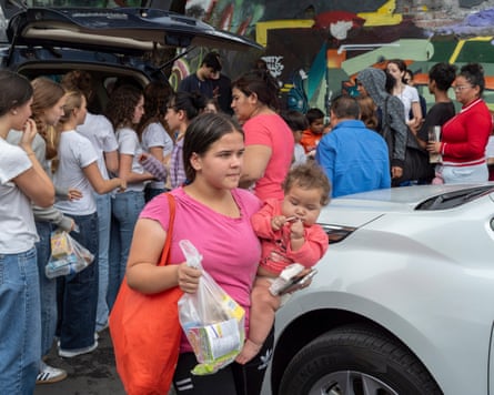 A woman holds a baby on one arm and a bag of food in the other hand. People queue behind her.