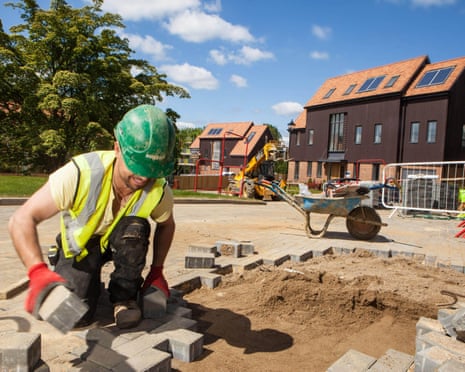 A construction worker in a green hard hat and high-visibility vest lays paving blocks on a sandy construction site
