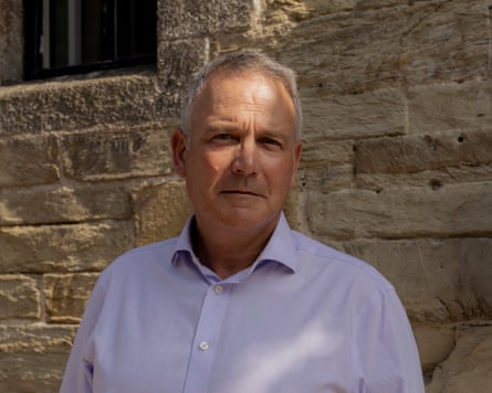 Head and shoulders image of Mike Bird wearing an open-necked pale-lilac shirt, standing against a brick wall, looking at the camera