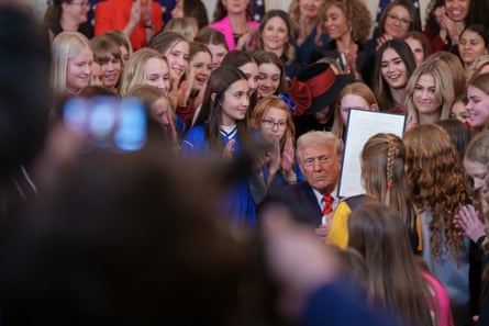 Donald Trump holds up a piece of paper, He is surrounded by women in sports uniforms.