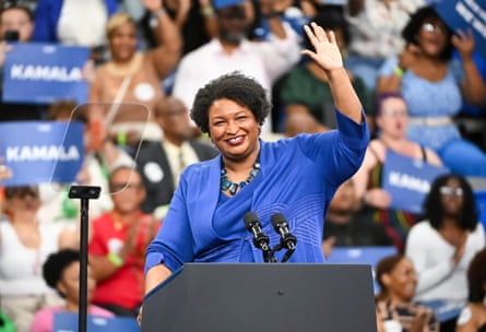 woman wearing blue stands at podium and waves with crowd of people behind her