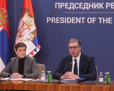 Serbian president Vučić speaks at a table beside Ana Brnabić before flags and a blue backdrop