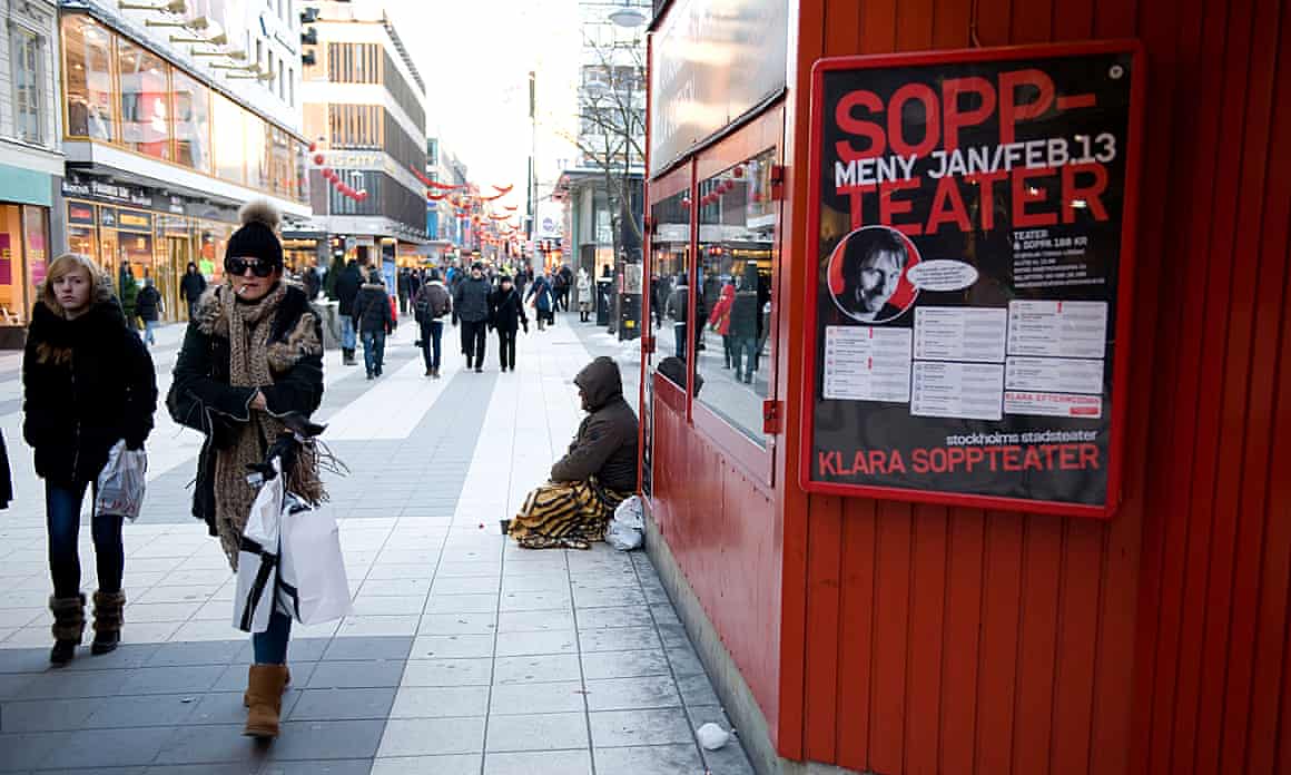Shoppers in winter clothing walk past a person sitting on the pavement outside a theatre in Stockholm, Sweden.