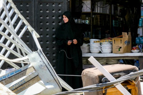 A woman in a black hijab and black dress stands in the midst of rubble under a bright sun.
