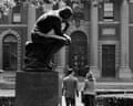 students walk past a statue of a man kneeling and holding his chin on his fist on a pedestal on a college campus