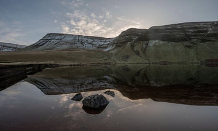 Reflections of mountains in a lake, in the middle of which sits a large rock, in Llyn Y Fan Fach, Carmarthenshire, Wales.