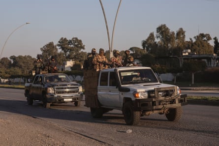 Armed men stand in the flatbeds of pickup trucks which are being driven down a wide road