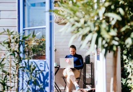 A woman in a blue jumper sits working on a laptop in a blue garden shed.