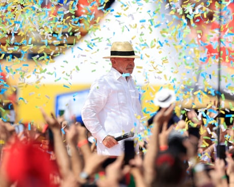 Lula, wearing a white shirt and a hat, holds a microphone as he walks along a stage surrounded by blue, green and yellow confetti and supporters cheering for him