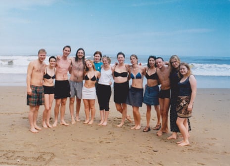 13 young people in swimming gear, standing in a row on a beach with the sea behind them