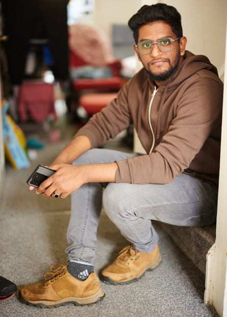 Akhil Jenny: he is photographed sitting at the bottom of a flight of stairs resting his feet on a grey carpet. He is a young Indian man with black hair and a short beard; he wears glasses, a brown zip-up hoodie, grey jeans and tan walking shoes.